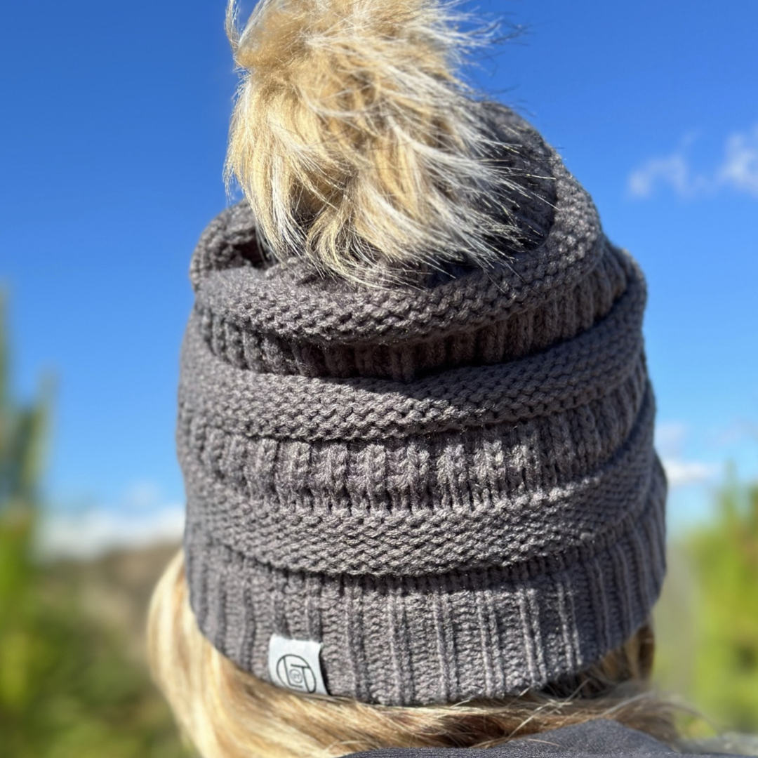 Someone is pictured from behind wearing the Women's Pom Beanie 2.0 in charcoal by Life Out There Apparel, complete with a fluffy pom-pom on top. The background displays a clear blue sky and blurred greenery.
