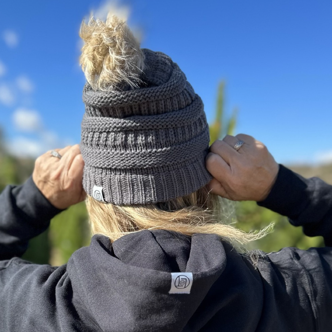 A person wearing the Life Out There Apparel Women's Pom Beanie 2.0 in charcoal stands outdoors on a sunny day. They are adjusting their hat against a blurred background of trees and a bright blue sky.