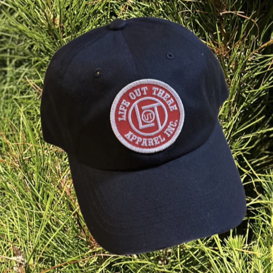 The LOT Snapback "Dad Hat" Trucker Hat in black, featuring a red and white circular patch with the Life Out There Apparel logo, is stylishly displayed against green pine needles. This adjustable cap ensures a comfortable fit for everyone.