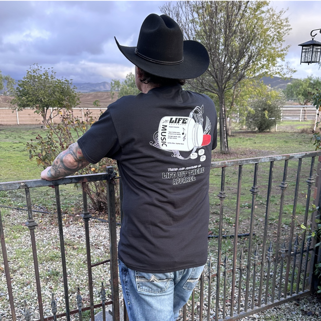 Life Out There Apparel “Music Is Medicine “ Black Unisex tee shirt is a medicine pill bottle tipped over with pills that have fallen out, that have music notes on them. This photo has a man wearing a cowboy hat showing the back of the shirt, looking out onto a ranch.
