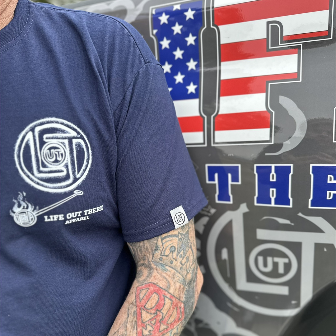 Wearing a navy Unisex Branded T Shirt from Life Out There Apparel, a person stands next to a vehicle decorated with a partial American flag design and eye-catching letters, while their tattooed arm is prominently visible in the foreground.