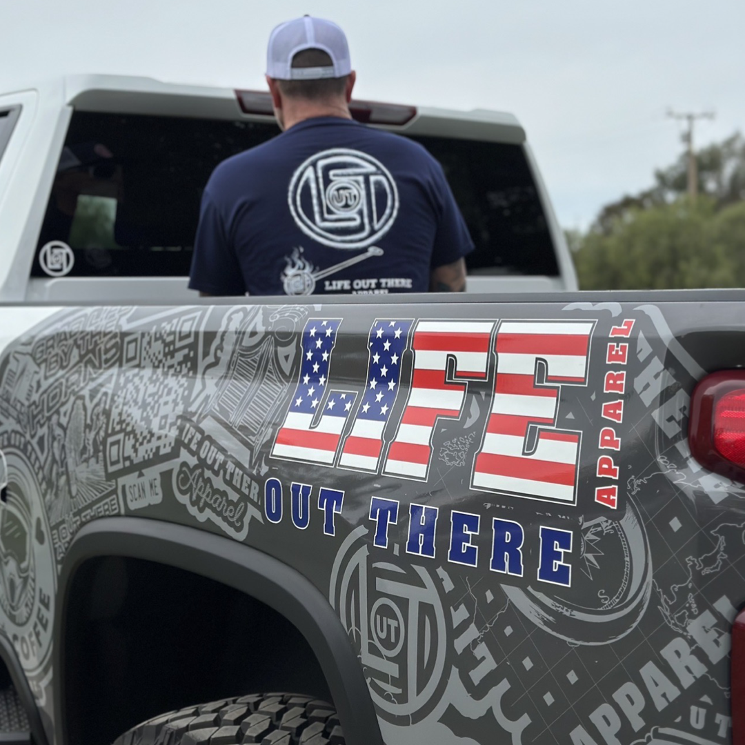A person wearing a Life Out There Apparel unisex branded t-shirt in navy stands by a white truck with a decorated tailgate. The tailgate features the Life Out There Apparel branding in patriotic colors, alongside various logo designs and stickers.