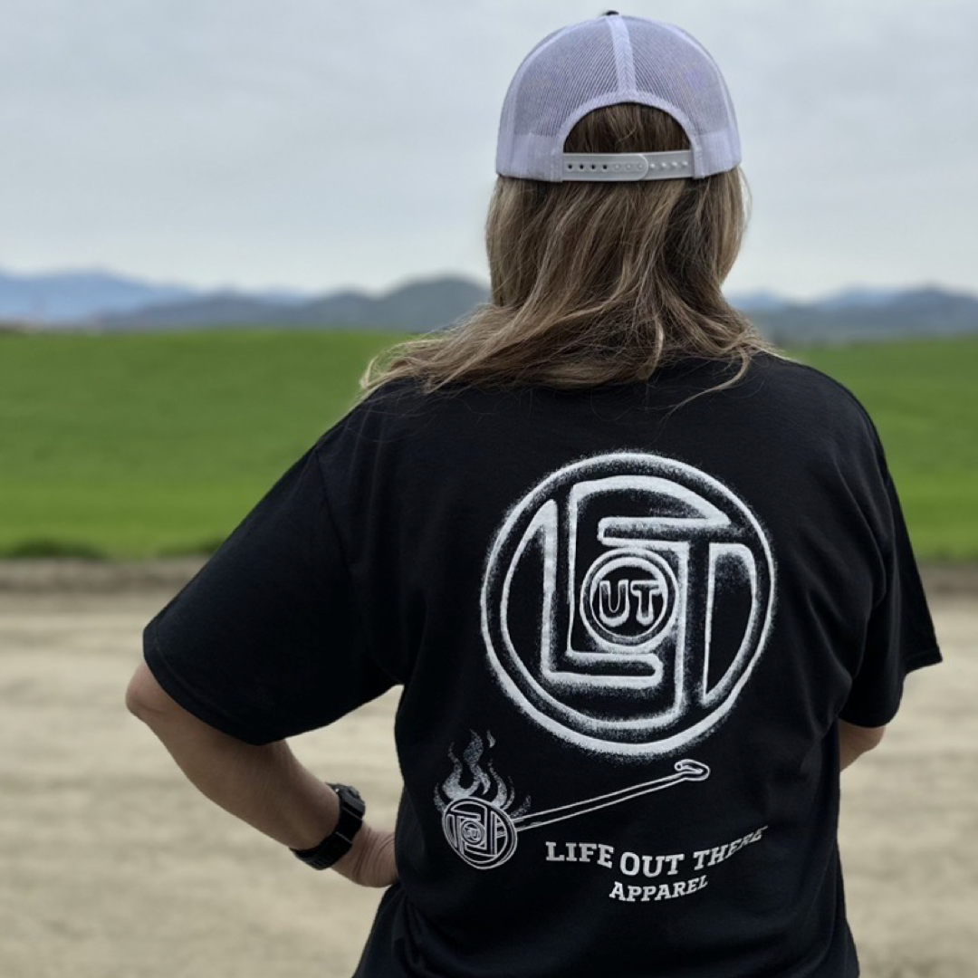 Life Out There Apparel “Branded” Unisex black tee shirt is a round logo of the companies initials, made with a hot branding iron. This is a photo of a women wearing the shirt, showing the back, with a field in front of her