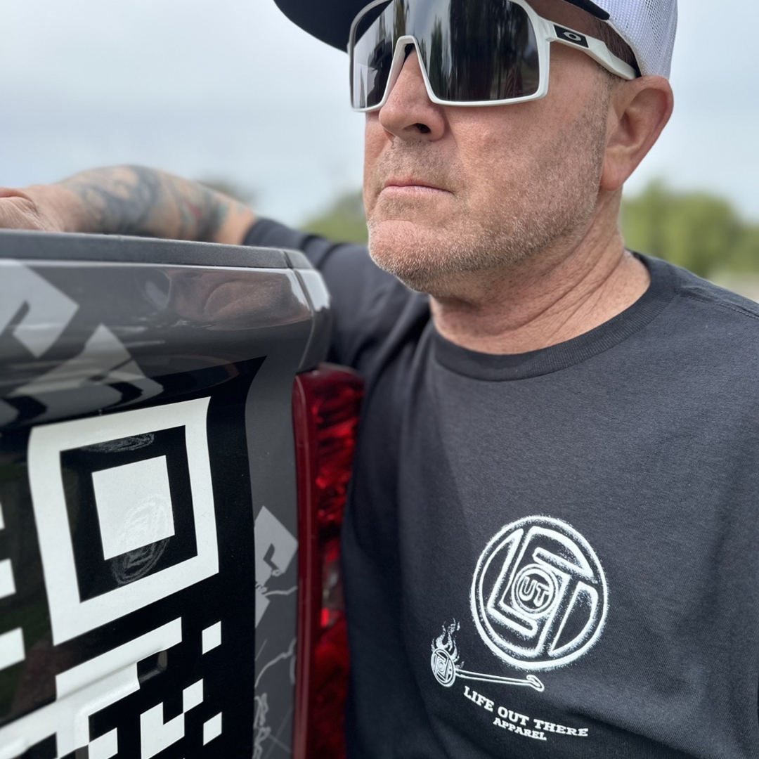Life Out There Apparel “Branded” Unisex black tee shirt is a round logo of the companies initials, made with a hot branding iron. This is a photo of a man wearing the shirt leaning up, against his truck.