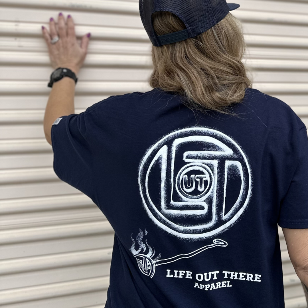 A person wearing a navy Unisex Branded T Shirt from Life Out There Apparel, along with a black cap, stands facing a beige corrugated metal wall. Showcasing the brand's logo recognition, the shirt is adorned with a circular emblem and a smoking pipe design.
