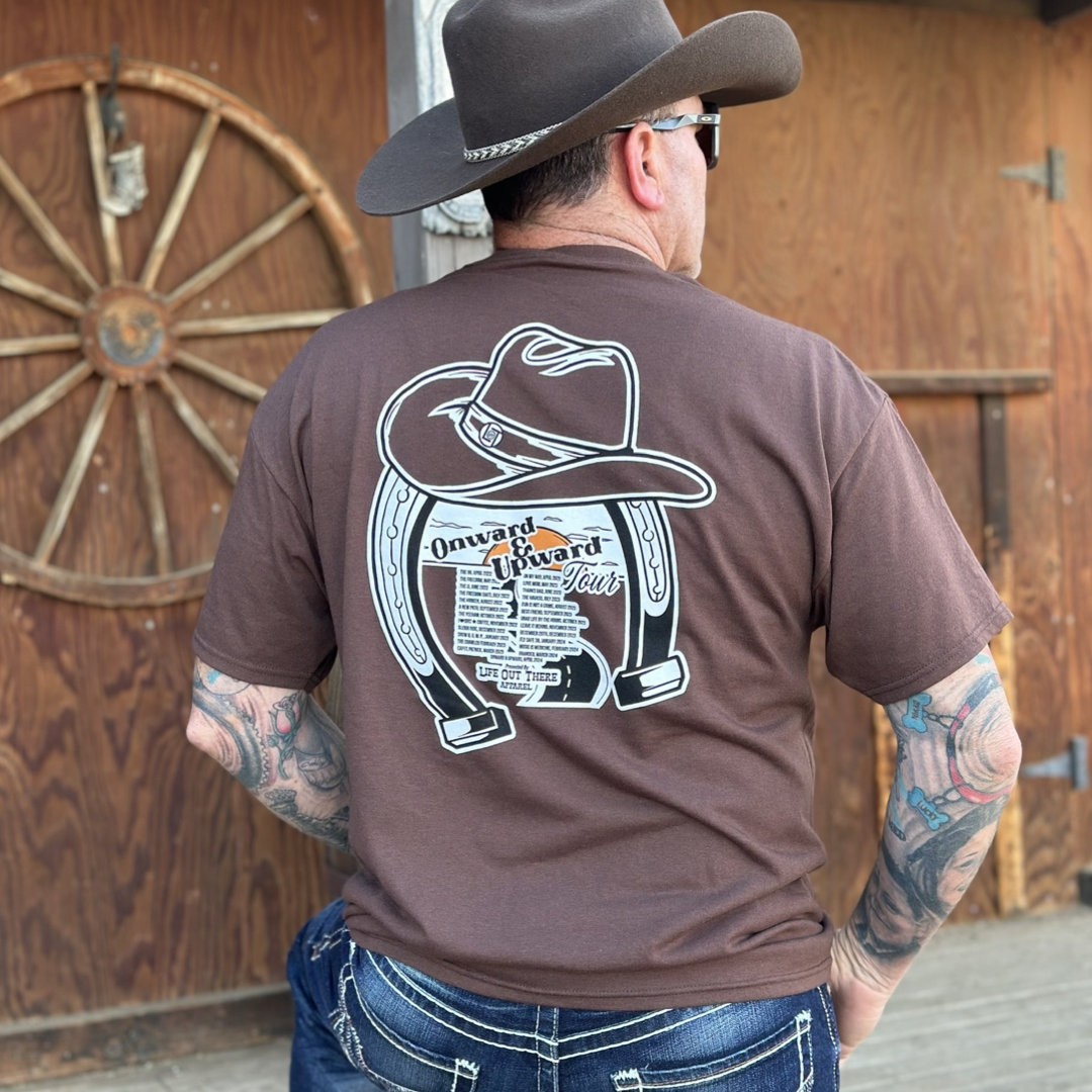 “ONWARD & UPWARD” Tour :
The Chocolate Brown Life Out There Apparel “ONWARD & UPWARD” Tour Unisex tee is a country concert design with a cowboy hat & horseshoe on the front and back, with all the design names from the past 2 years (instead of city & tours dates) This photo is of a man wearing a cowboy hat standing in front of a tack / feed room on a ranch.