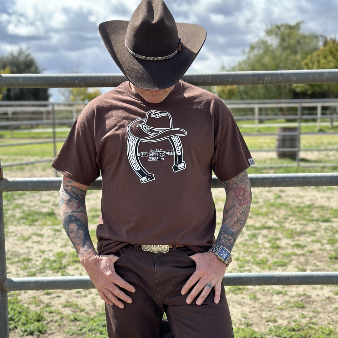 “ONWARD & UPWARD” Tour :
The Life Out There Apparel “ONWARD & UPWARD” Tour Unisex tee in Chocolate Brown is a country concert design with a cowboy hat & horseshoe on the front and back, with all the design names from the past 2 years (instead of city & tours dates) This photo is of a man wearing a cowboy hat standing on a ranch next to a round pin.