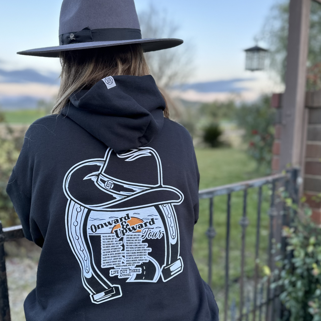 Life Out There Apparel “ONWARD & UPWARD” Tour Black Unisex hoodie is a country concert design with a cowboy hat & horseshoe on the front and back with every Life Out There Apparel launch for the past 2 years. This photo shows a woman wearing a cowboy hat looking out into a ranch.