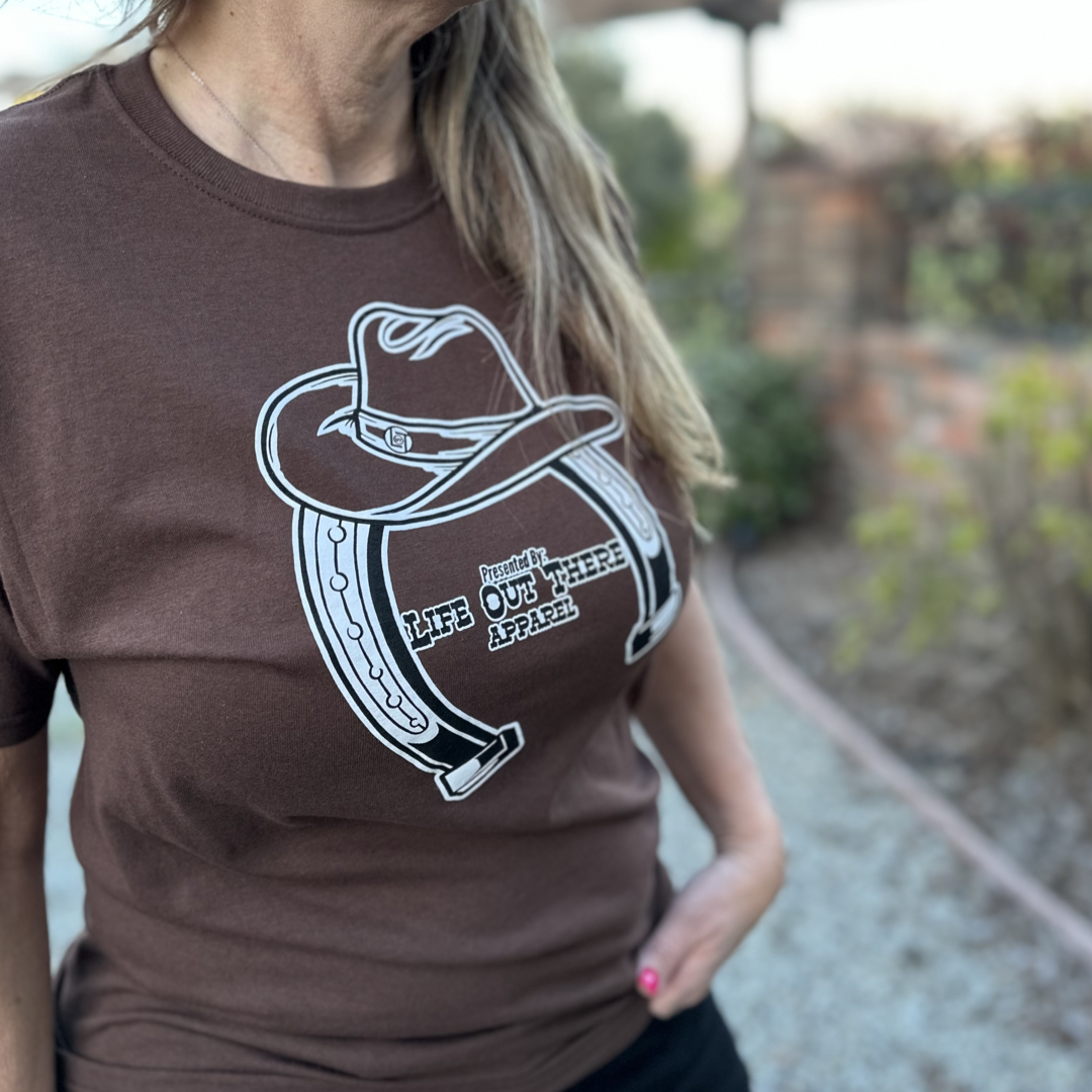 “ONWARD & UPWARD” Tour :
The Chocolate Brown Life Out There Apparel “ONWARD & UPWARD” Tour Unisex tee in is a country concert design with a cowboy hat & horseshoe on the front and back, with all the design names from the past 2 years (instead of city & tours dates) This photo is of a woman wearing a cowgirl standing on a ranch.