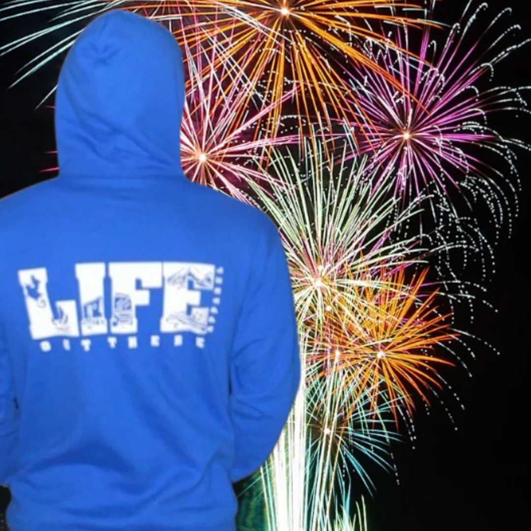 Someone wearing the Unisex The OG Hoodie in Royal/White from Life Out There Apparel stands against a backdrop of colorful fireworks illuminating the night sky, evoking memories of camping in the desert where stars create their own fiery display.