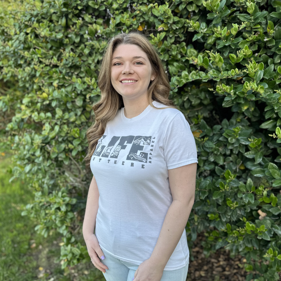 A woman with long brown hair, dressed in The OG Women's T Shirt - White/Grey by Life Out There Apparel and light jeans, stands smiling in front of lush green bushes, envisioning her next dirt bike adventure on a sunny day.