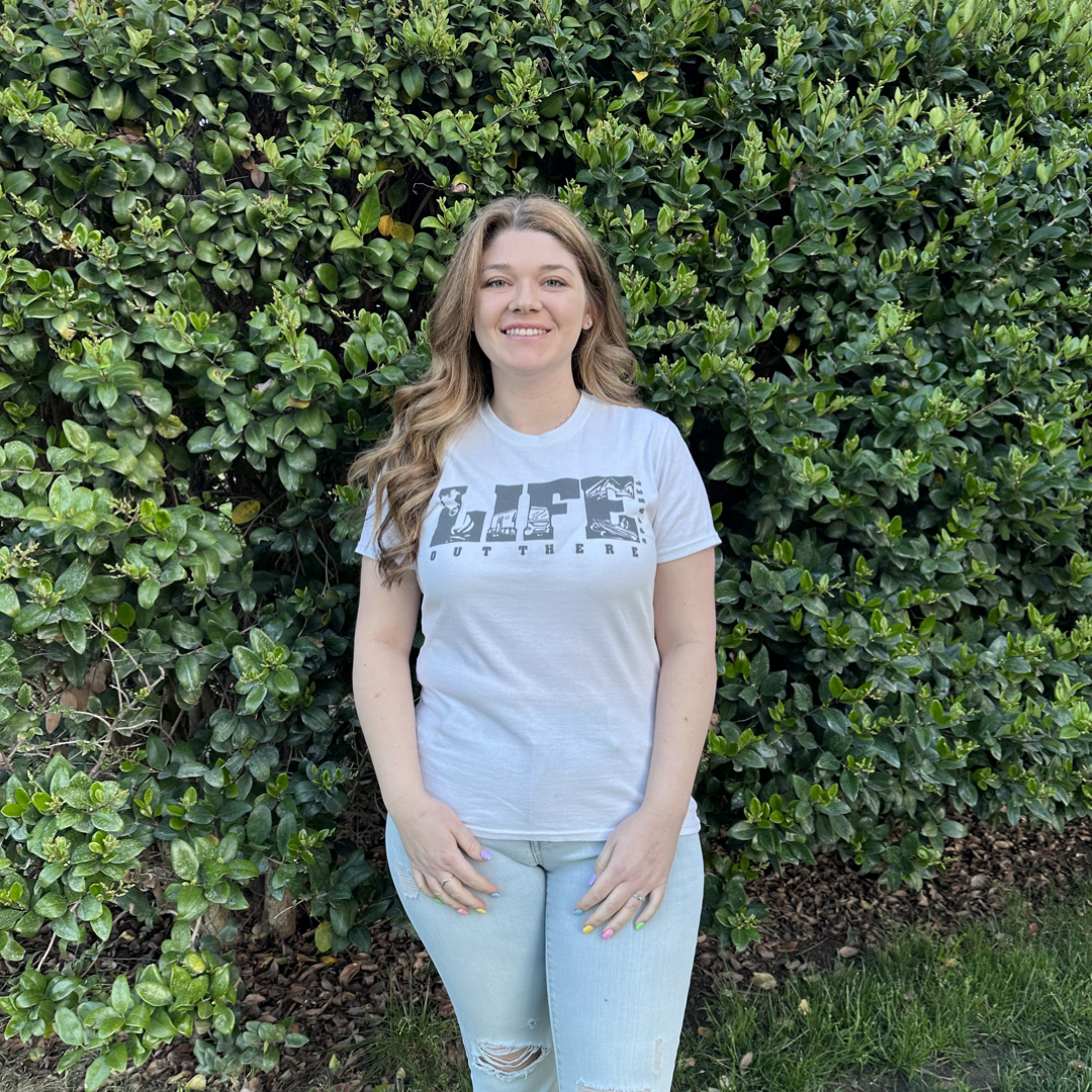 A woman stands smiling in front of a lush green hedge, wearing The OG Women's T Shirt - White/Grey by Life Out There Apparel and light blue jeans, her casual style reminiscent of weekend camping adventures.