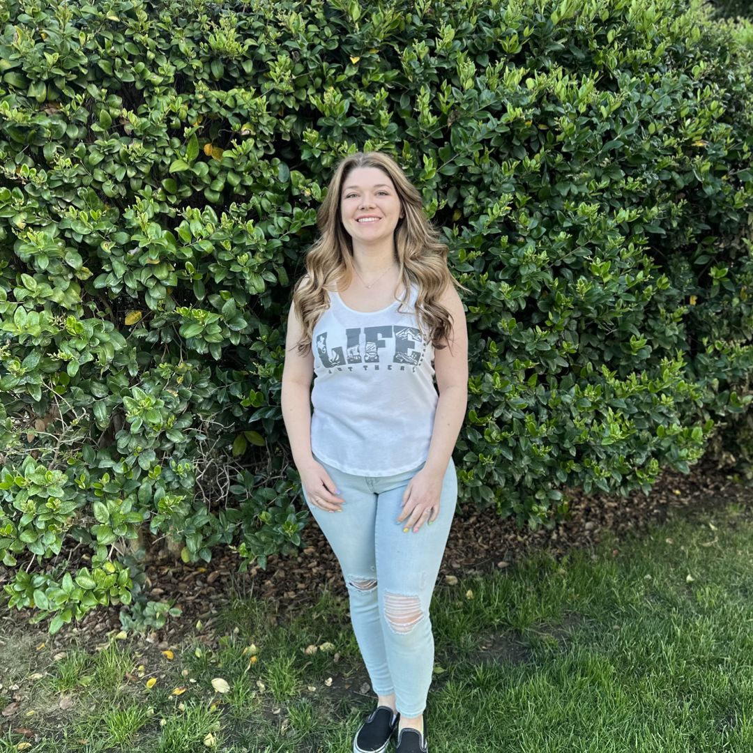 A woman with long brown hair stands smiling in front of a large, lush green hedge. Wearing The OG Women's Tank Top in White/Grey from Life Out There Apparel, light blue ripped jeans, and black shoes, she looks ready for an adventure. The grassy ground with patches of brown leaves hints at her love for camping under the stars.