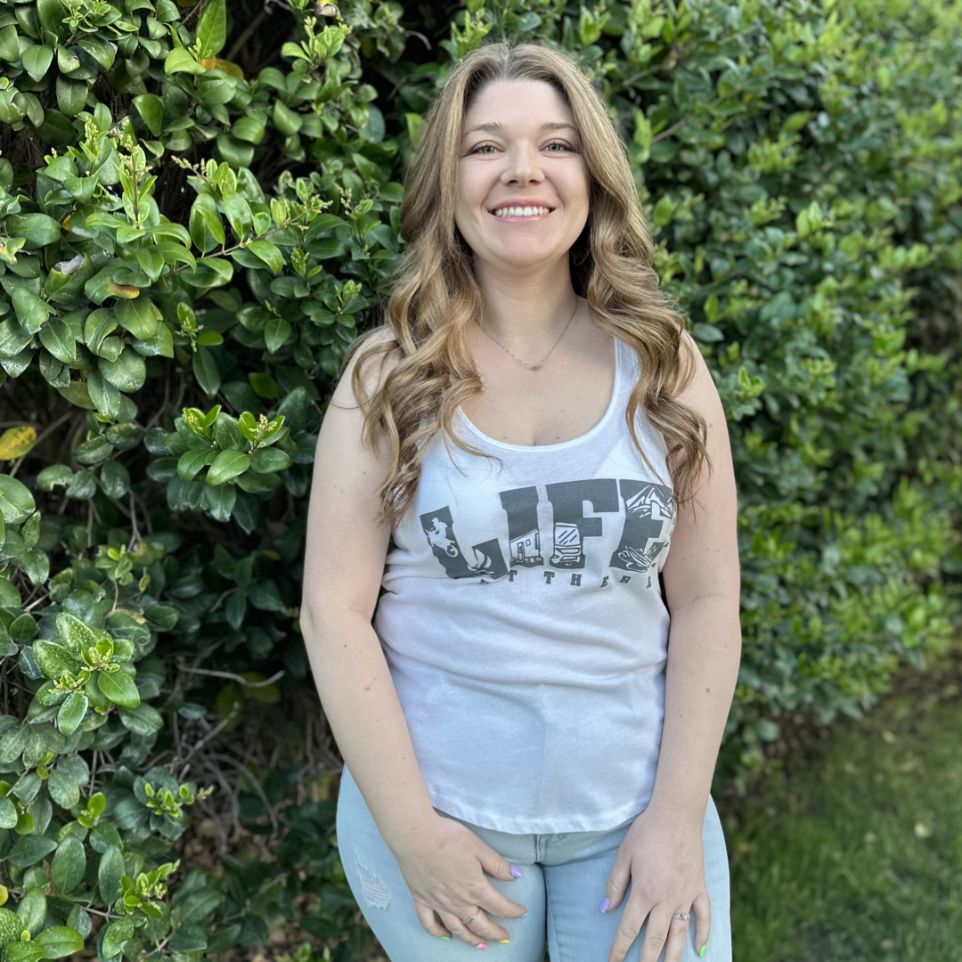 A woman with long, curly hair and a big smile stands outdoors in front of lush green shrubs. She wears Life Out There Apparel's The OG Women's Tank Top in White/Grey along with light blue jeans, embodying the adventurous spirit of camping.