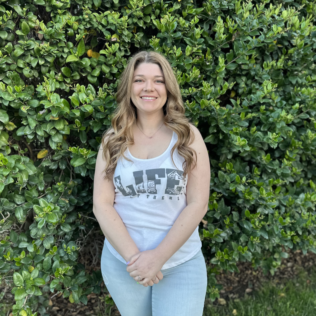 A woman with long, wavy hair stands smiling in front of a lush green hedge as if she's just returned from an exhilarating weekend of dirt biking. She is wearing The OG Women's Tank Top in White/Grey from Life Out There Apparel and light blue jeans, with her hands clasped in front.