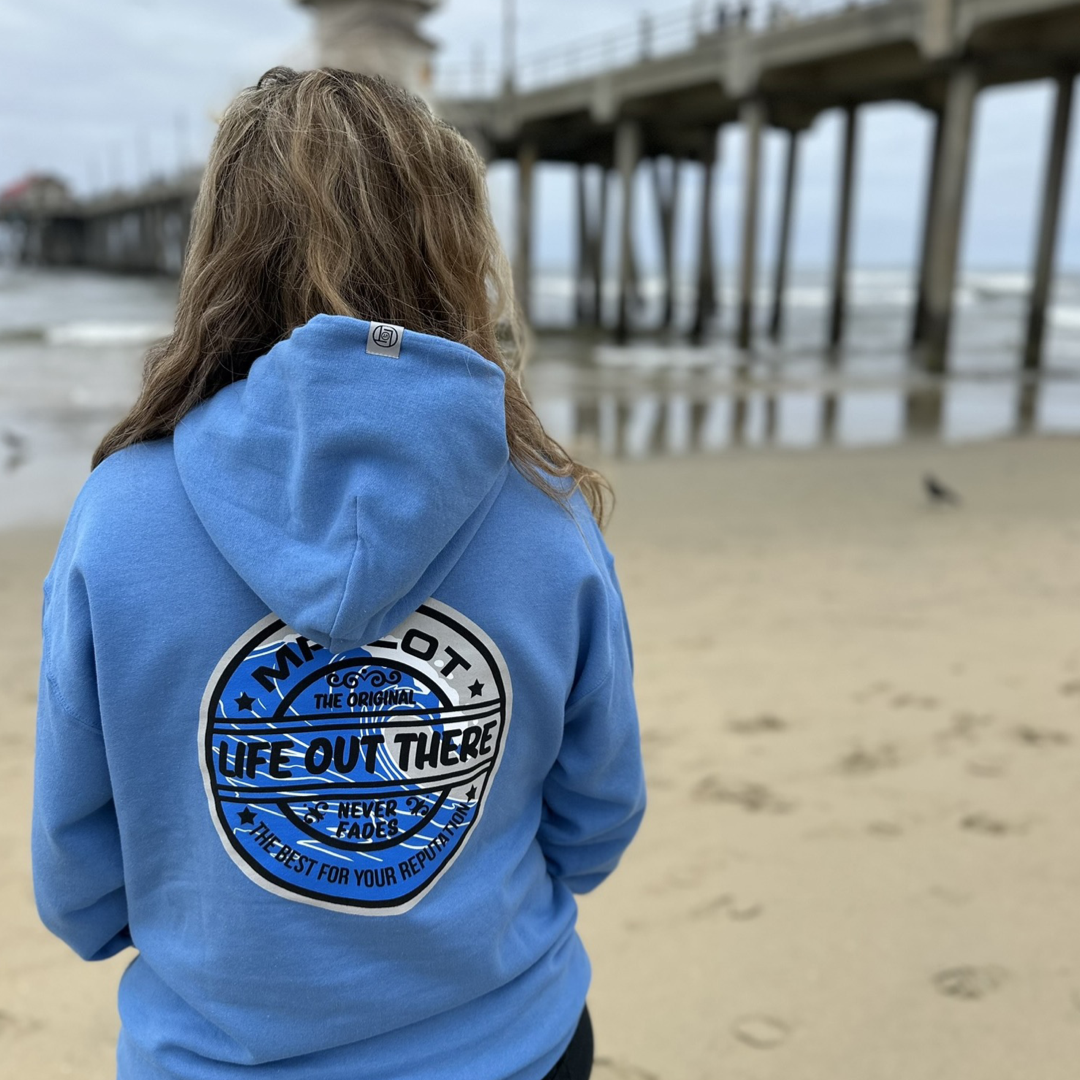 Wearing a Columbia Blue Unisex Summer Vibes Hoodie from Life Out There Apparel, a person stands on the sandy beach, gazing at the ocean with "Life Out There, Never Fade" emblazoned on their back. The pier stretches over the water as summer vibes linger in the overcast sky.