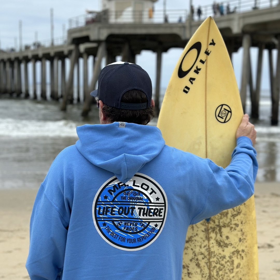A person wearing the Unisex Summer Vibes Hoodie in Columbia Blue and a cap stands on a beach, the coconut scent of surfboard wax lingering as they hold a yellow surfboard with an Oakley logo. Facing the ocean near a pier under a cloudy sky, their hoodie from Life Out There Apparel reads LIFE OUT THERE, capturing those ultimate SUMMER VIBES.