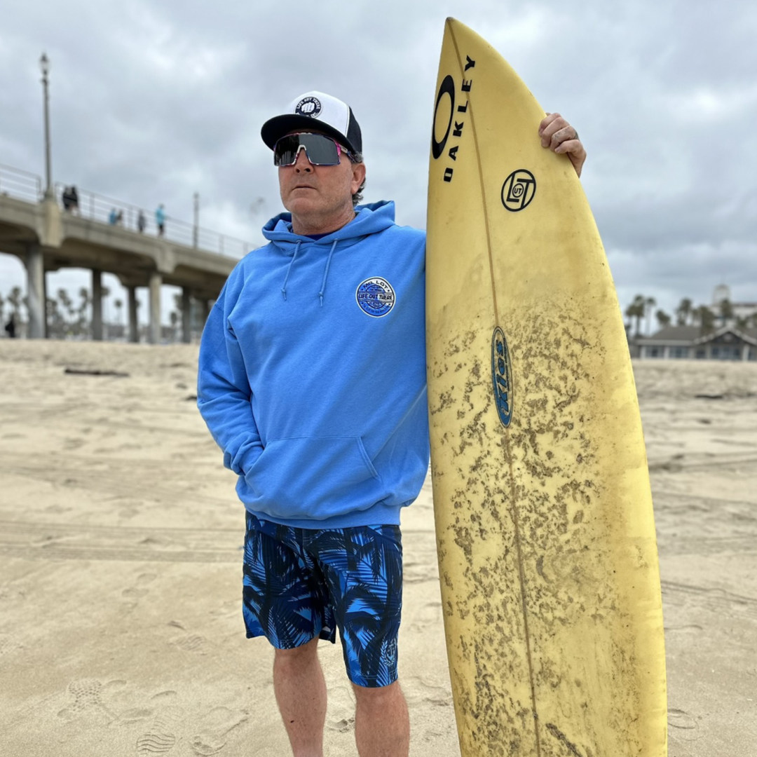 Wearing a Life Out There Apparel Unisex Summer Vibes Hoodie in Columbia Blue and palm-patterned shorts, a person stands on the beach, exuding summer vibes while holding a yellow surfboard upright. They accessorize with a cap and sunglasses, with surfboard wax nearby as the pier and cloudy sky create the perfect backdrop.