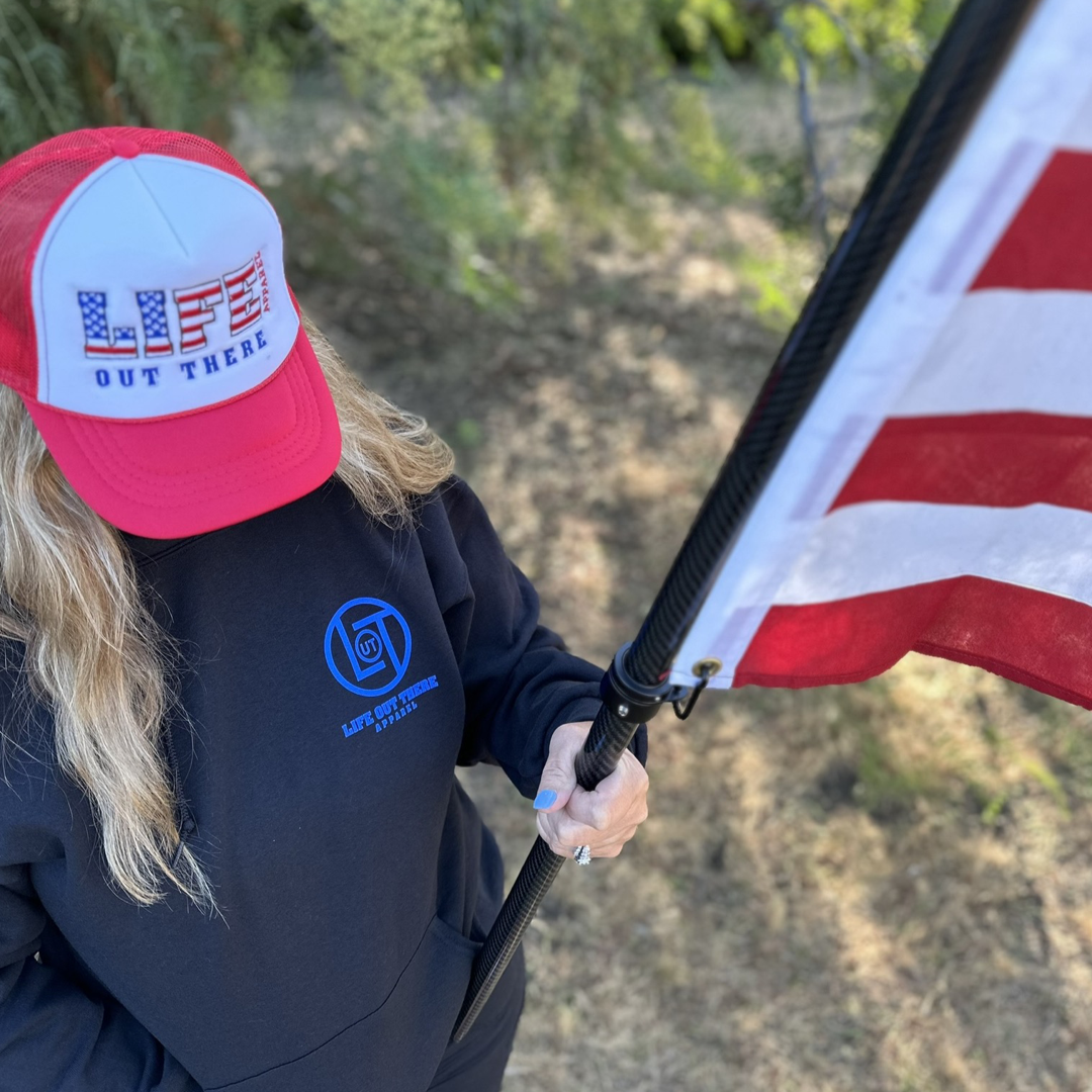 A person with long blonde hair wearing a Life Out There Apparel cap and a Unisex God Bless America Hoodie in black holds an American flag upright. The background of trees and grass sets a patriotic scene, echoing the spirit of the 4th of July in the United States of America.
