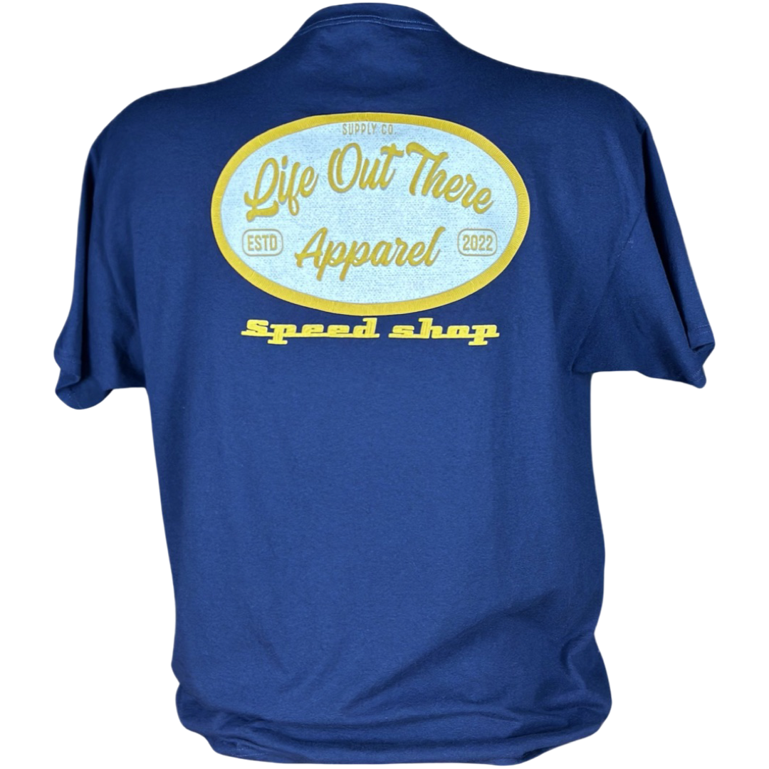 A navy t-shirt from Life Out There Apparel showcases an embroidered patch on the back with a yellow and white oval emblem. The design includes "ESTD 2022," "SUPPLY CO," and prominently features "Speed Shop" in bold yellow text, paying homage to the automotive industry.
