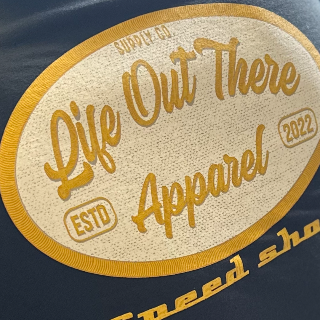 Close-up of a black Unisex Speed Shop T-Shirt from Life Out There Apparel, featuring an embroidered patch. The patch displays the text 'Life Out There Apparel', alongside 'SUPPLY CO', 'ESTD 2022', and gold-toned decorative elements inspired by classic Speed Shop designs.