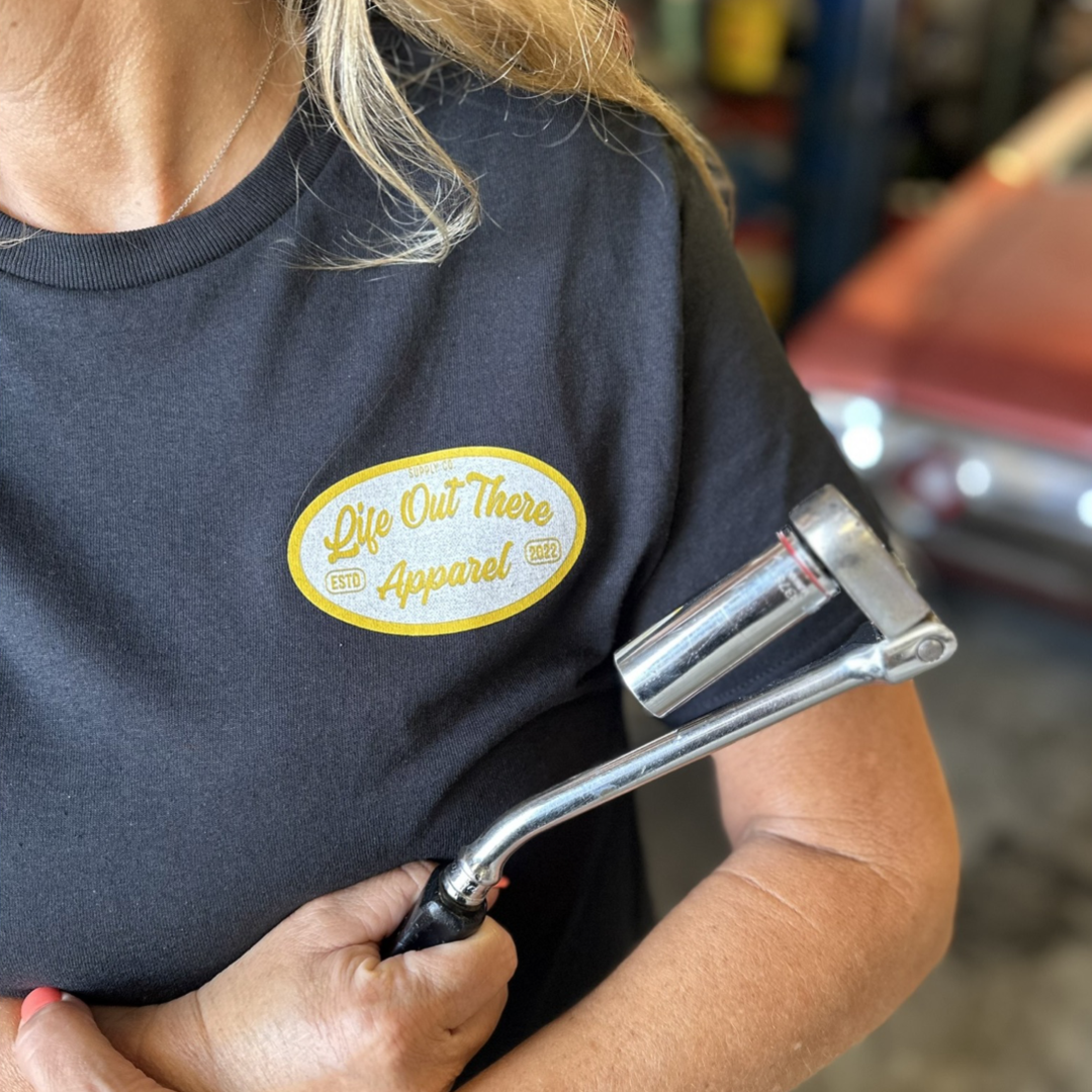 A person holding a tire iron is wearing an Unisex Speed Shop T Shirt - Black from Life Out There Apparel, featuring an embroidered patch. In the background, a vintage car, awaiting its next custom modification, is softly blurred.
