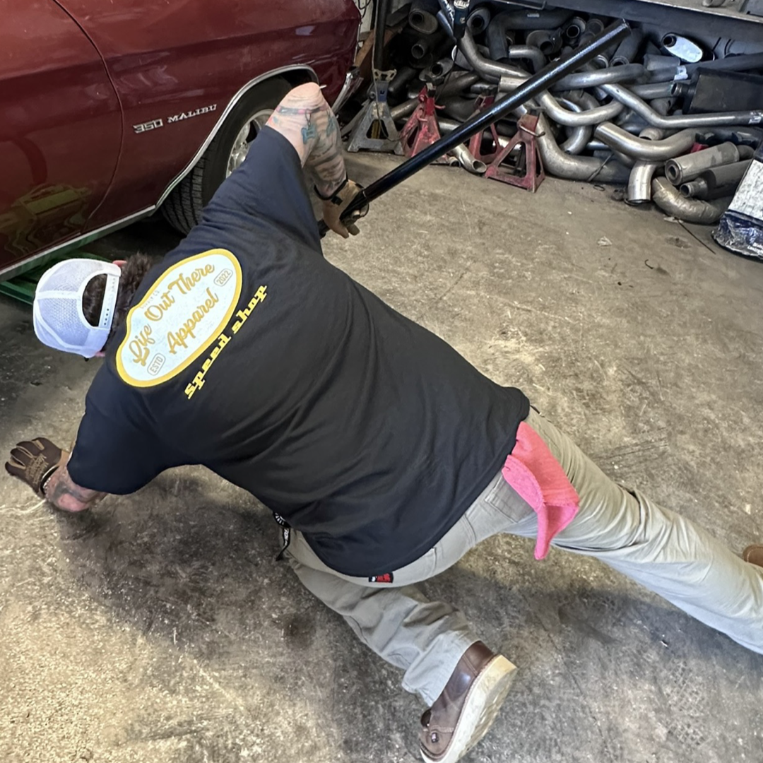 Dressed in a Unisex Speed Shop T-Shirt in black from Life Out There Apparel and beige pants adorned with embroidered patches, the person works on a car's undercarriage in a garage. They're lying on the floor, reaching under a red vehicle, surrounded by various automotive parts and tools from the car customization scene.