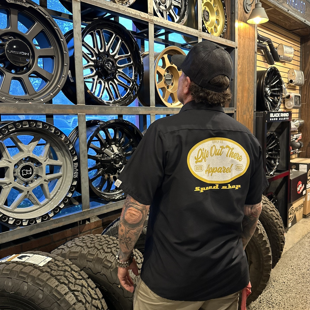 A person wearing a Life Out There Apparel's Unisex Speed Shop Collard Button Up Shirt in black inspects a display of custom car rims mounted on the wall. The shelves, filled with various styles and sizes, perfectly capture the essence of the automotive industry in its rustic, tire-laden interior.