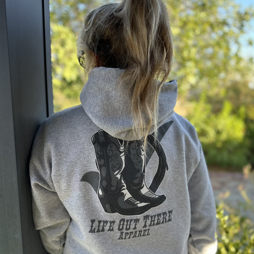 A person with long hair stands near a window, wearing the Unisex Boot Check Hoodie in Oxford Grey from Life Out There Apparel. Outside, vivid green foliage symbolizes the vibrancy of life during Suicide Awareness and Prevention Month.