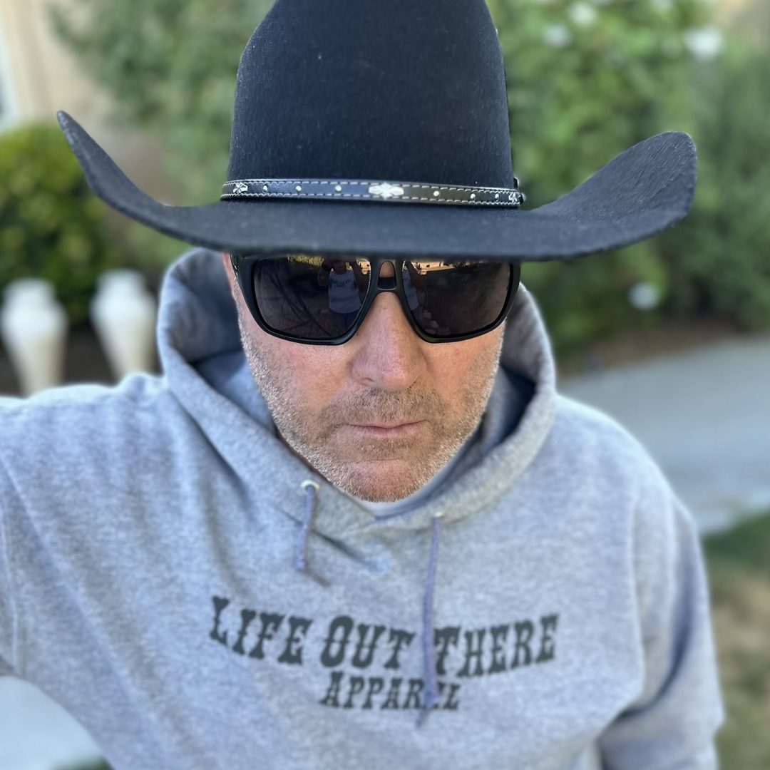 A person wearing a black cowboy hat, dark sunglasses, and an Oxford Grey Unisex Boot Check Hoodie from Life Out There Apparel stands outdoors during suicide awareness month. The background is slightly blurred with greenery.