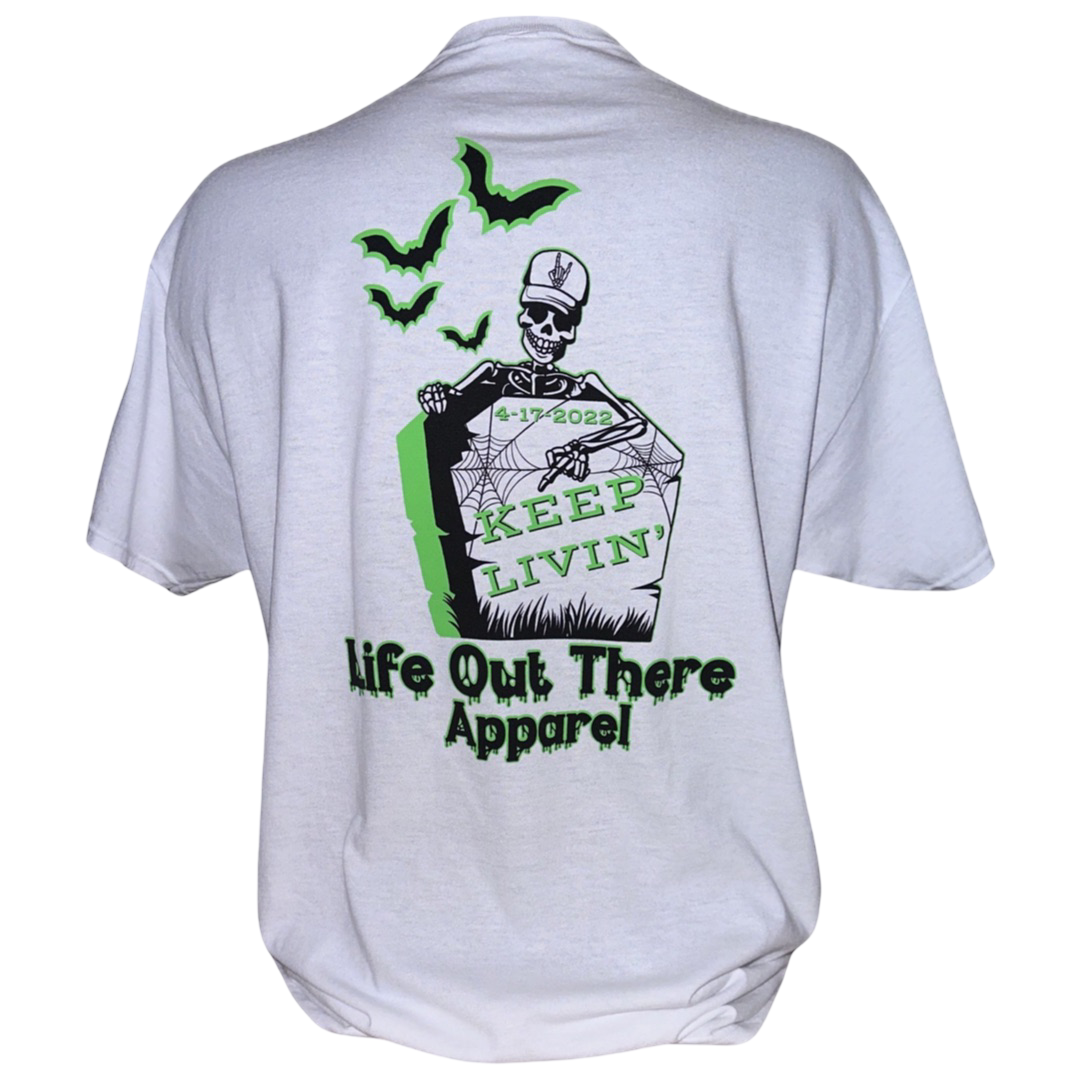 Unisex Keep Livin' T Shirt - White by Life Out There Apparel features a skeleton emerging from a coffin with the date 4-17-2022 and the phrase "Keep Livin." Flying bats enhance the Halloween atmosphere, while "Life Out There Apparel" is printed below in green and black—a perfect holiday favorite for all skeleton enthusiasts.