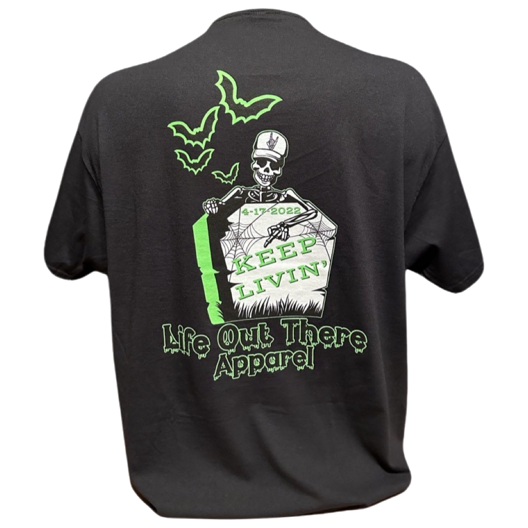 Unisex Keep Livin' T Shirt - Black from Life Out There Apparel showcases a spooky skeleton gripping a gravestone, embellished with "Keep Livin" and the date 4-17-2022 in neon green. Ideal for Halloween, it features two hovering bats and spine-chilling font below.