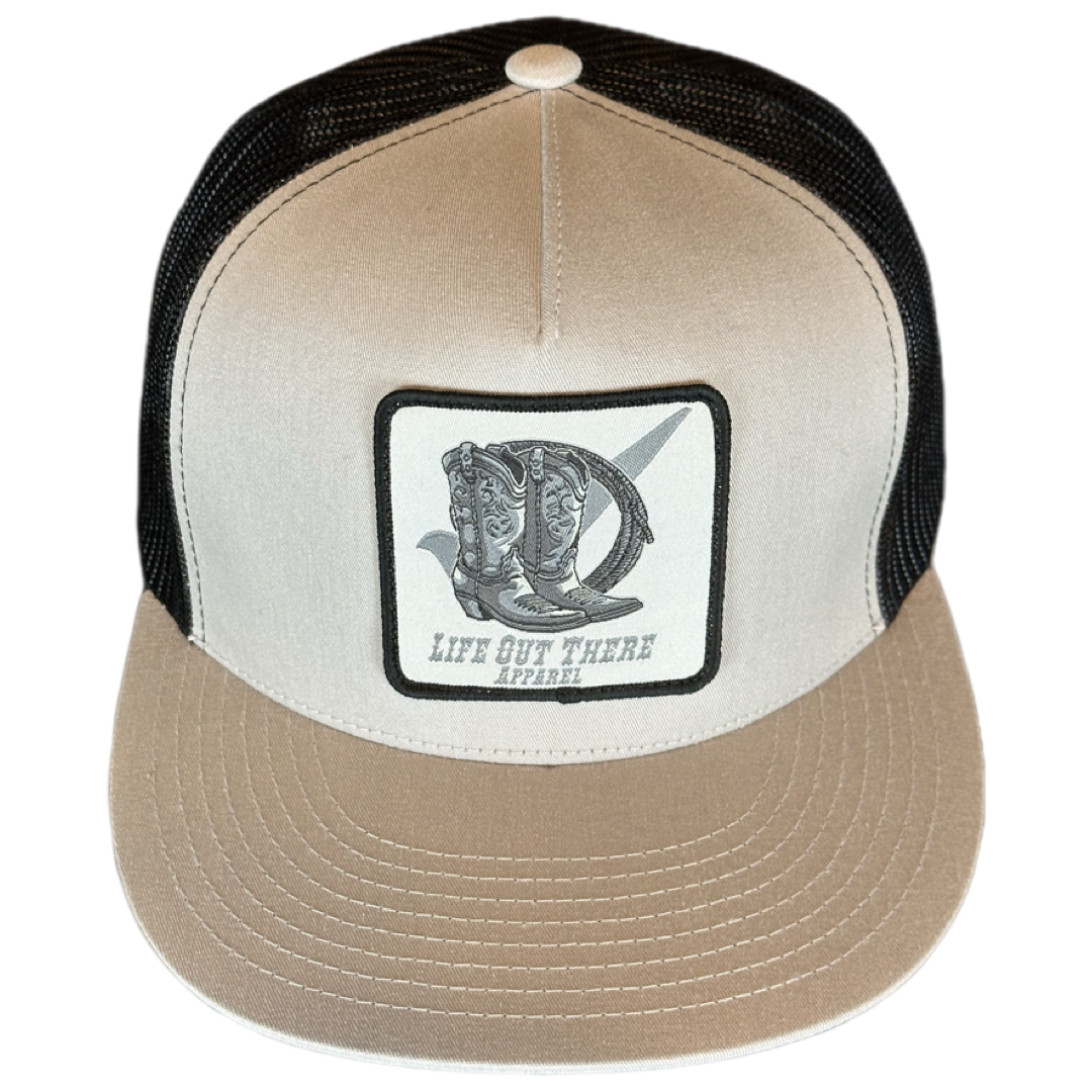 A silver and black BOOT CHECK Snapback Trucker Hat by Life Out There Apparel, featuring a flat bill brim with visible stitching. It includes a stitched patch on the front illustrating a cowboy boot with a spur and the text "LIFE OUT THERE APPAREL" in bold letters.