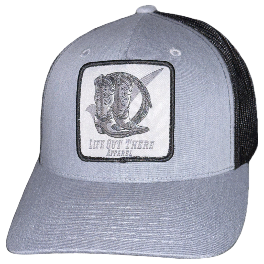 The BOOT CHECK Curved Bill Snapback Trucker Hat in Heather Grey/Black highlights a patch adorned with an illustration of cowboy boots and a hat, and proudly features the brand name "Life Out There Apparel.