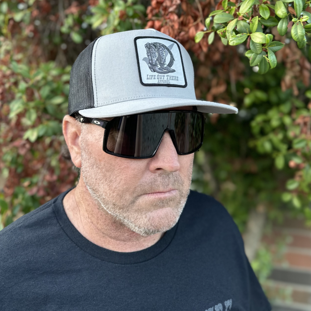 A man wearing the BOOT CHECK - Flat Bill Snapback Trucker Hat from Life Out There Apparel, in silver and black, and large sunglasses stands in front of a leafy background, looking to the side. He's dressed in a black shirt and has light facial hair.