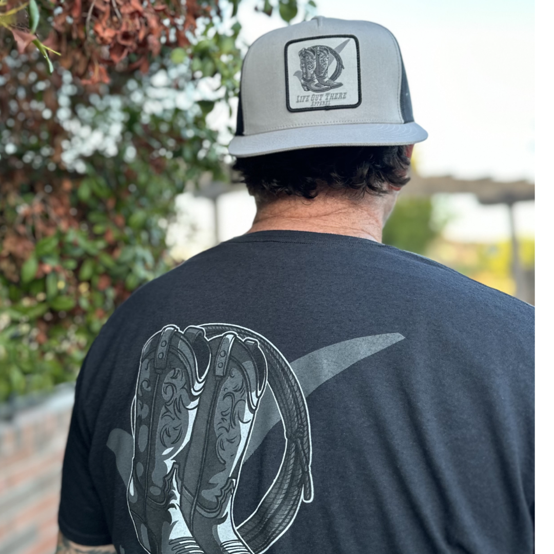 A man is wearing the Life Out There Apparel BOOT CHECK - Flat Bill Snapback Trucker Hat in silver and black, along with a black t-shirt featuring a stylized cowboy boot design on the back. He is standing outdoors near some bushes, with blurred greenery and a brick wall in the background.