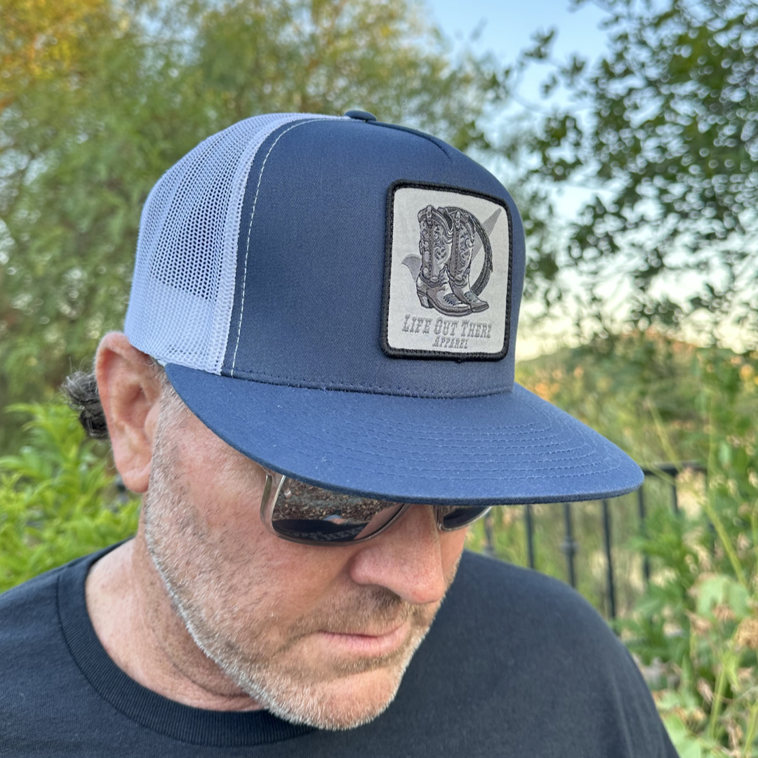 A man wearing the "BOOT CHECK - Flat Bill Snapback Trucker Hat" in navy and white from Life Out There Apparel stands outside. The sunny day showcases lush greenery and a tree-filled background, while his sunglasses add an extra touch of cool to the scene.