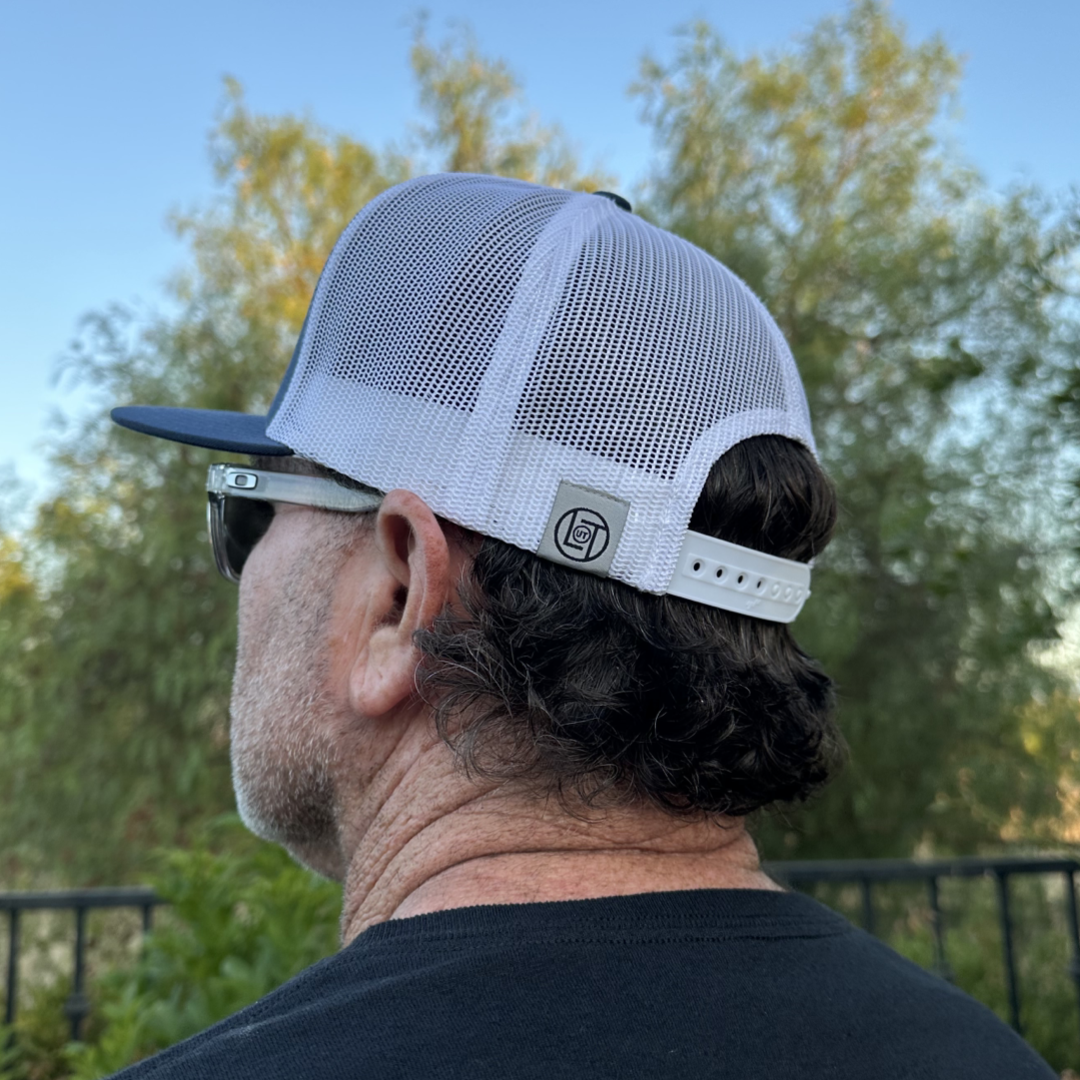 A man with curly hair is seen in profile, sporting the Life Out There Apparel BOOT CHECK - Flat Bill Snapback Trucker Hat in Navy/White worn backwards along with sunglasses. He stands outdoors on a sunny day surrounded by greenery, and a black metal fence can be seen behind him.