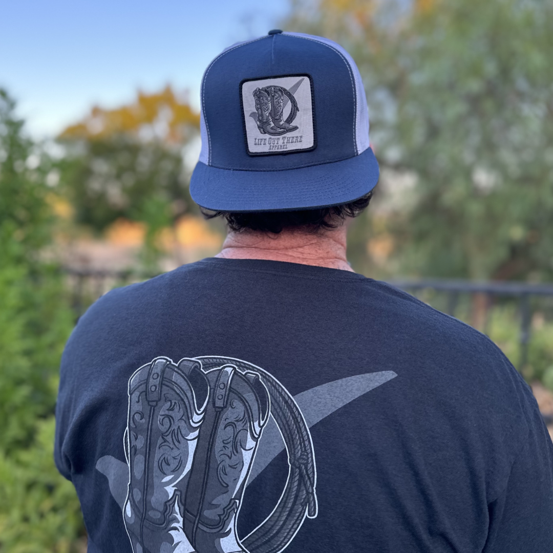 A person wearing a Life Out There Apparel BOOT CHECK Flat Bill Snapback Trucker Hat in navy and white stands outdoors. The back of their black t-shirt features a graphic of cowboy boots and a horseshoe, set against lush green foliage and trees in the background.