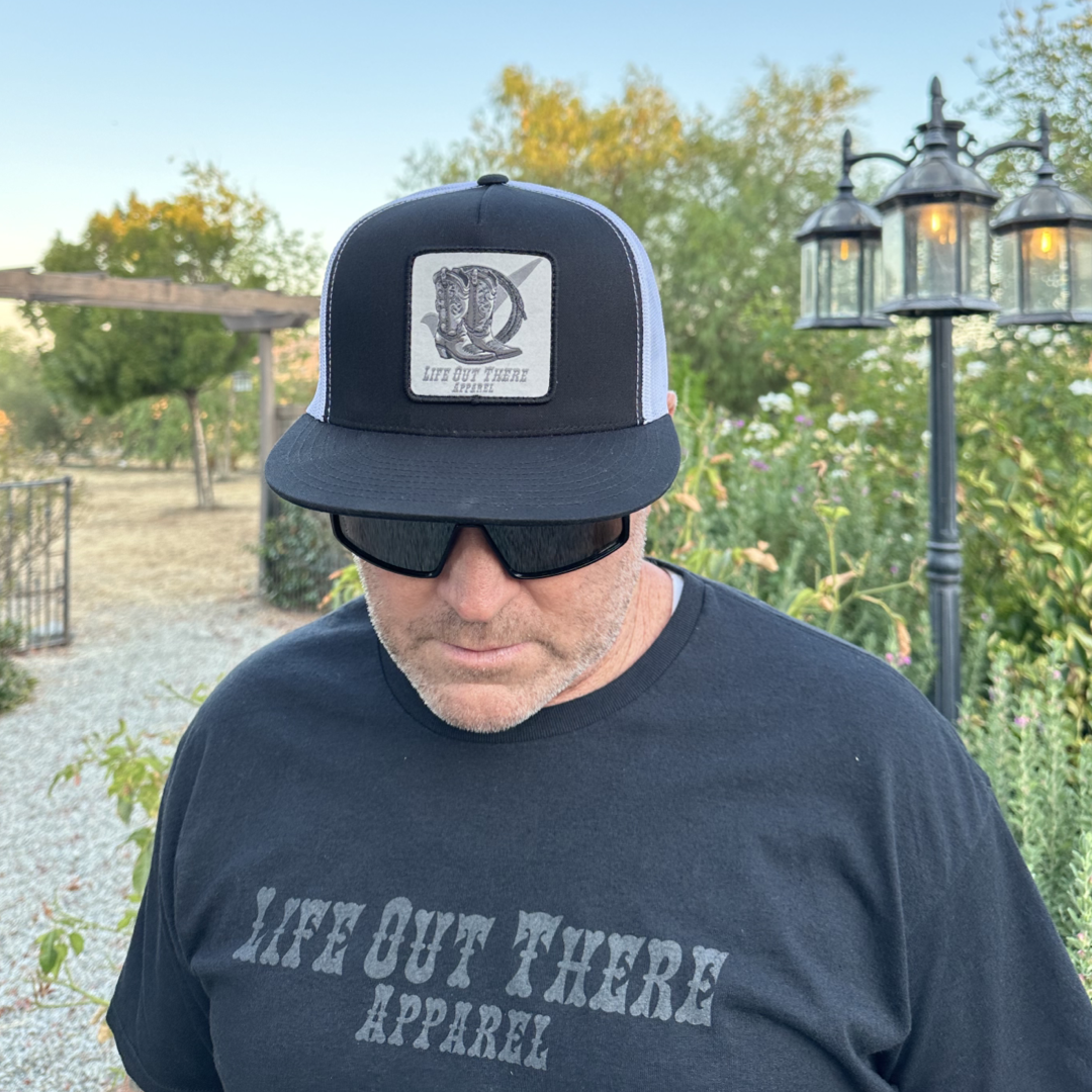 A man wearing a BOOT CHECK - Flat Bill SnapBack Trucker Hat in Black/White and sunglasses stands outdoors on a garden path surrounded by greenery. His shirt is by Life Out There Apparel, and an outdoor lantern casts light under the clear sky.