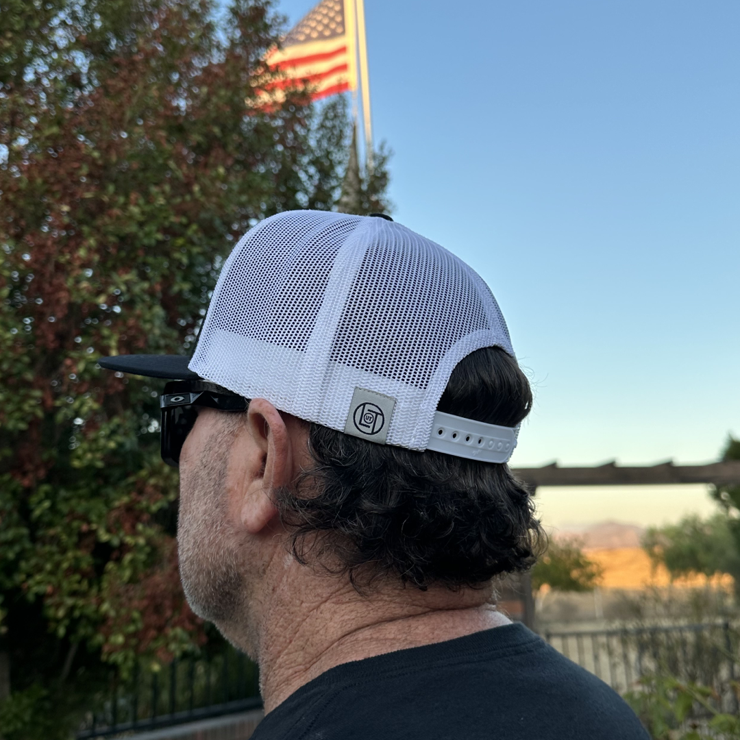 A man sporting a BOOT CHECK - Flat Bill SnapBack Trucker Hat in black and white from Life Out There Apparel and sunglasses gazes off into the distance. Behind him, an American flag flutters against the backdrop of a clear blue sky, with trees and a fence adding depth to the scenery.