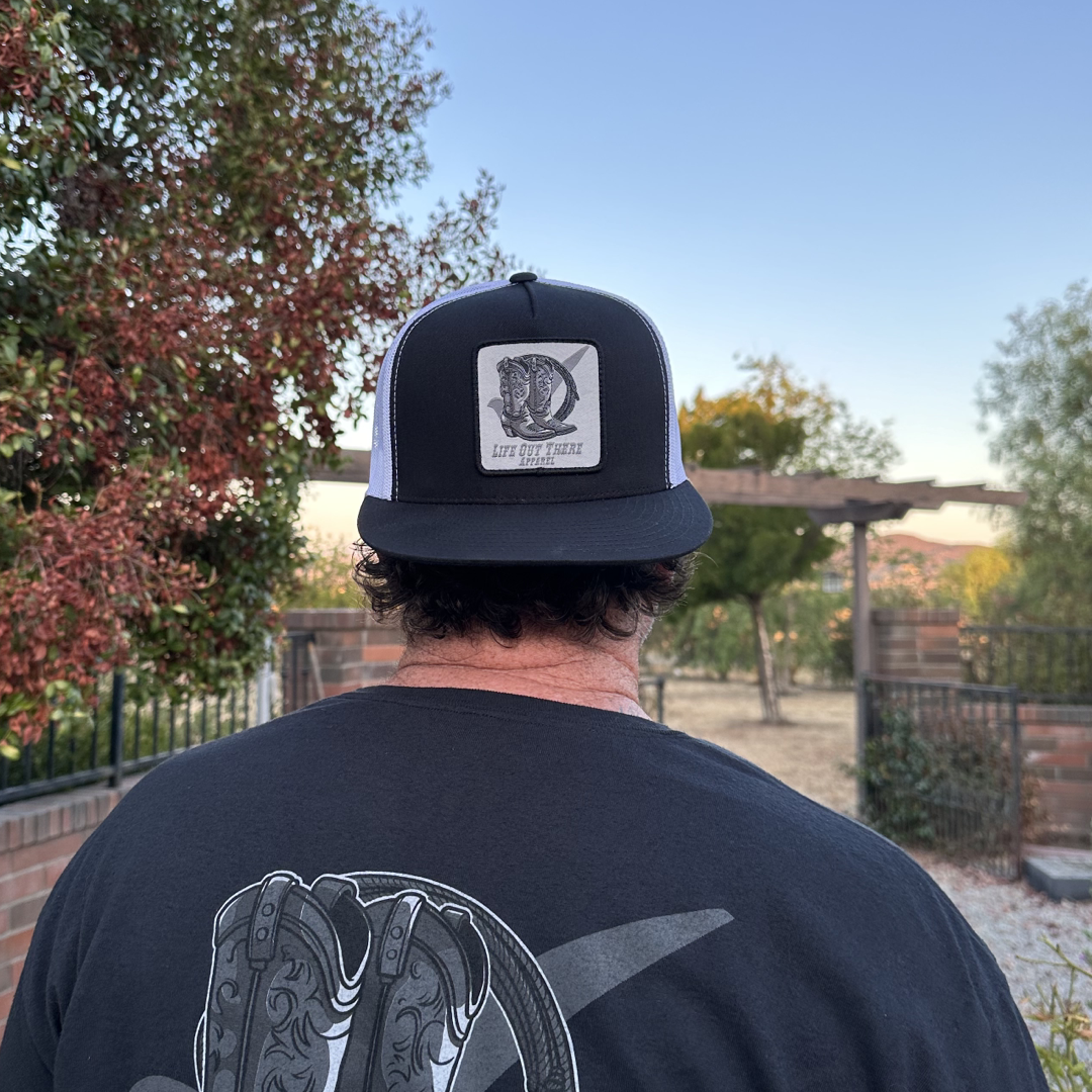Standing outdoors with dark hair, a person faces away wearing the BOOT CHECK - Flat Bill SnapBack Trucker Hat in black and white from Life Out There Apparel. They also sport a black T-shirt adorned with cowboy boots. The background showcases trees, a pergola, and a brick fence beneath the clear blue sky.