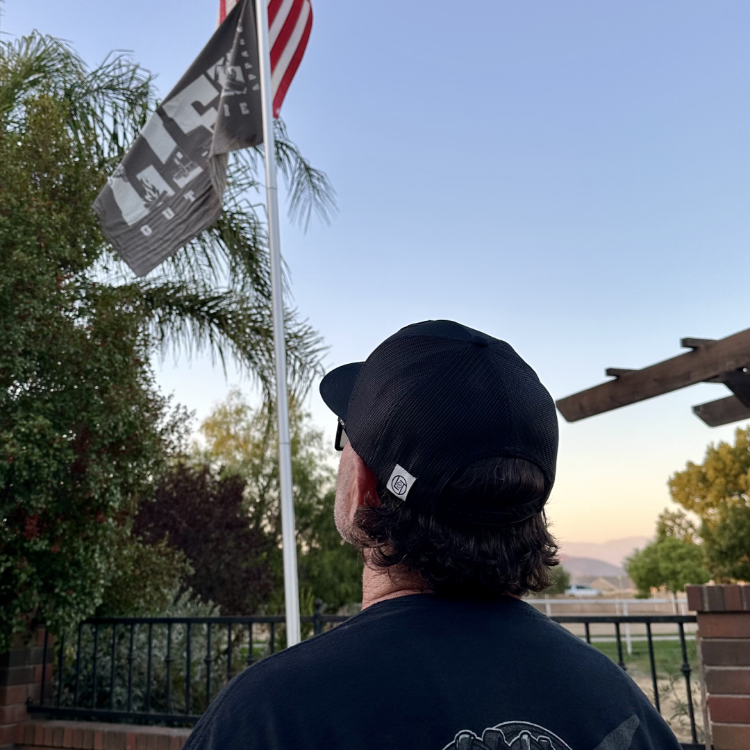 A person sporting a Life Out There Apparel BOOT CHECK - Flat Bill Snapback Trucker Hat in black and a matching shirt stands outdoors, admiring an American flag alongside another black flag on a flagpole. The scenic backdrop features trees, a fence, and a clear sky aglow with the warm hues of sunset.