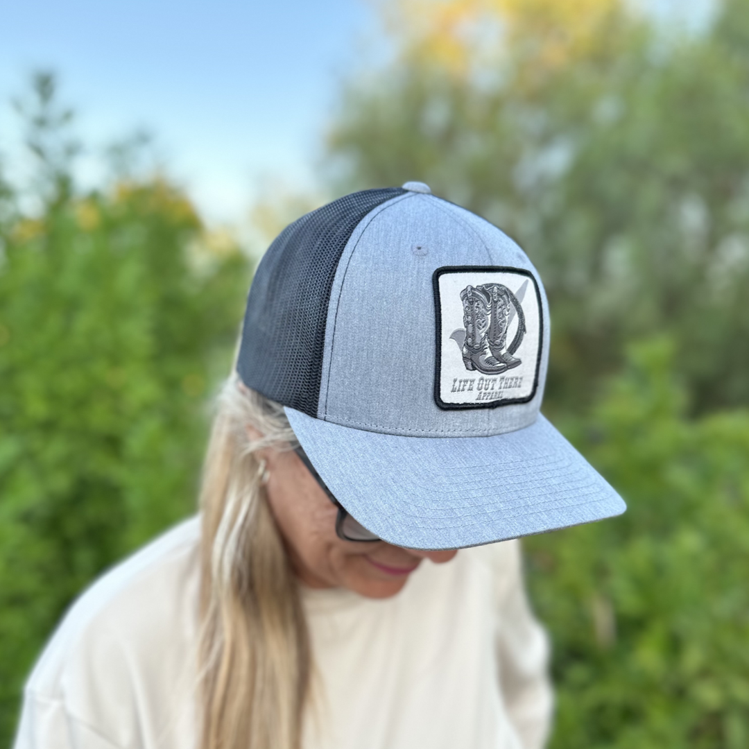 A person wearing the BOOT CHECK Curved Bill Snapback Trucker Hat in Heather Grey and Black from Life Out There Apparel, featuring a patch with an eagle and the text "Lite Out Here." The hat sits snugly on their head as they look down, revealing long hair. In the blurred background are green foliage and trees.