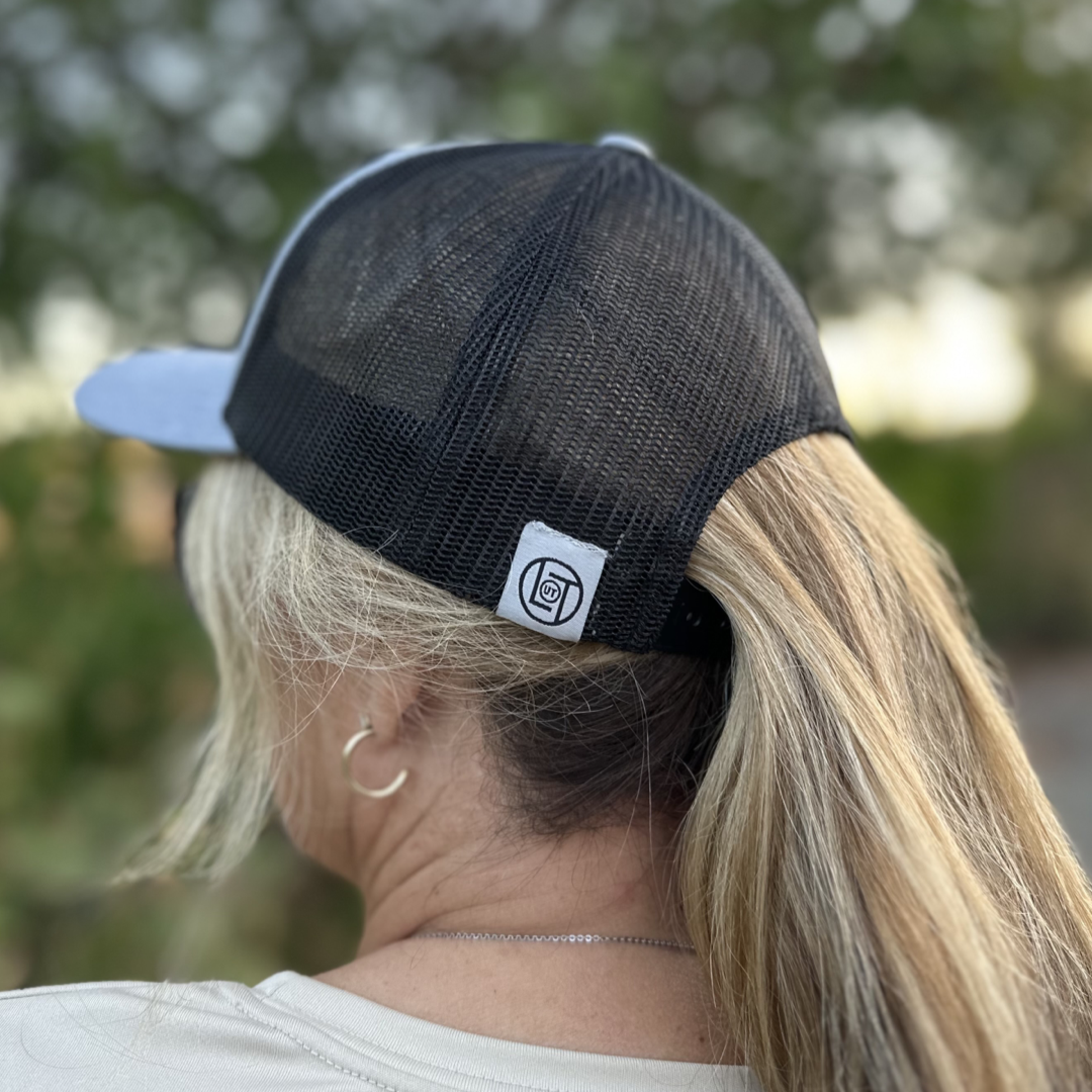 A person facing away from the camera sports a Heather Grey and Black BOOT CHECK - Curved Bill Snapback Trucker Hat by Life Out There Apparel, featuring a small logo on the back. Blonde hair is pulled through an opening in the hat's mesh design. They wear a light-colored shirt and a thin necklace, while green foliage creates a beautifully blurred background.