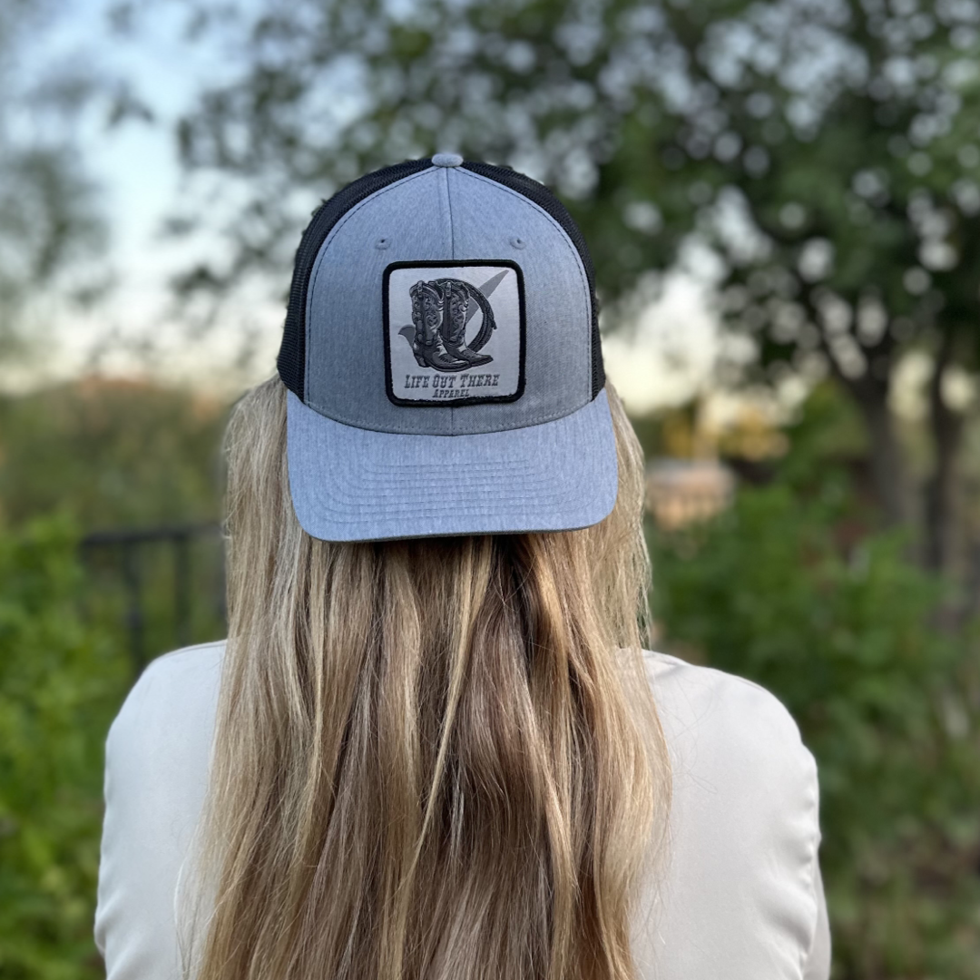 A person with long blonde hair is sporting the Life Out There Apparel's BOOT CHECK curved bill snapback trucker hat in heather grey and black, featuring a logo and the text LET OUR RANGE on the back. The stylish snapback perfectly complements their look as they stand outdoors, surrounded by trees and foliage in the backdrop.