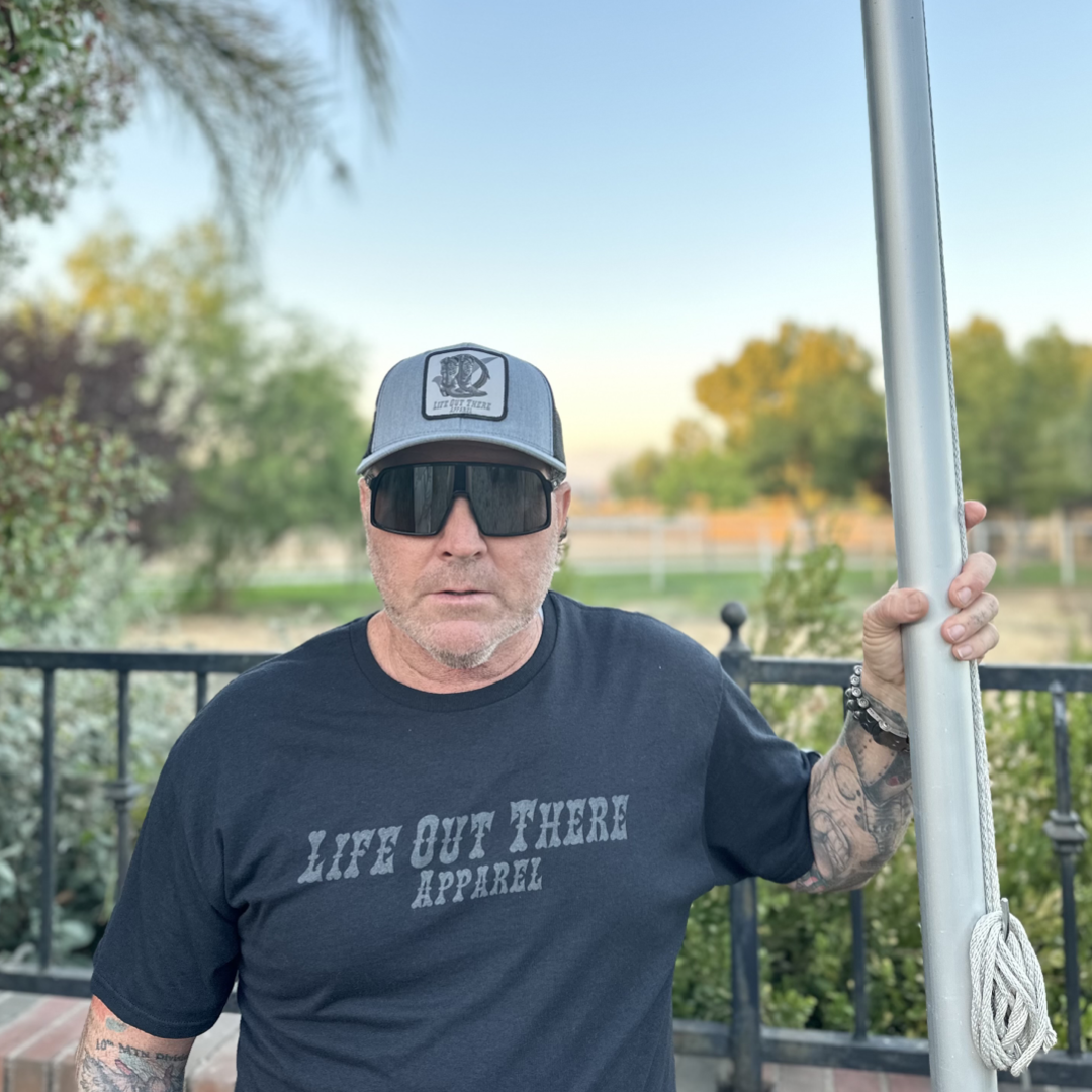 A man outdoors, wearing a black shirt and sunglasses, holds a pole while donning the BOOT CHECK Curved Bill Snapback Trucker Hat in Heather Grey and Black by Life Out There Apparel. Trees and a fence are visible in the background under the clear sky.