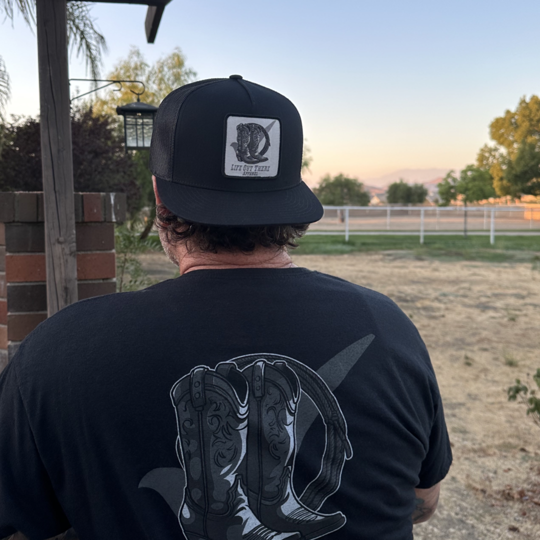 An individual sporting a black BOOT CHECK - Flat Bill Snapback Trucker Hat by Life Out There Apparel, along with a T-shirt featuring a boot design, stands outdoors beside a brick structure. In the background are dry grass, a white fence, trees, and a pastel sky at either dusk or dawn.
