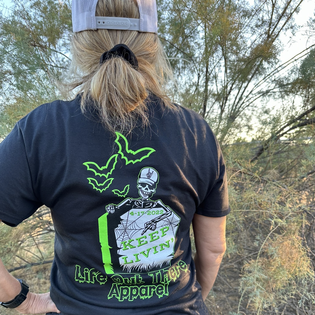 A person with blonde hair and a cap is facing away from the camera, wearing the Unisex Keep Livin' T Shirt in black by Life Out There Apparel. The T-shirt features a spooky neon green skeleton, bats, and tombstone design with the words "Keep Livin" visible. Bushes and trees provide a Halloween-esque backdrop.