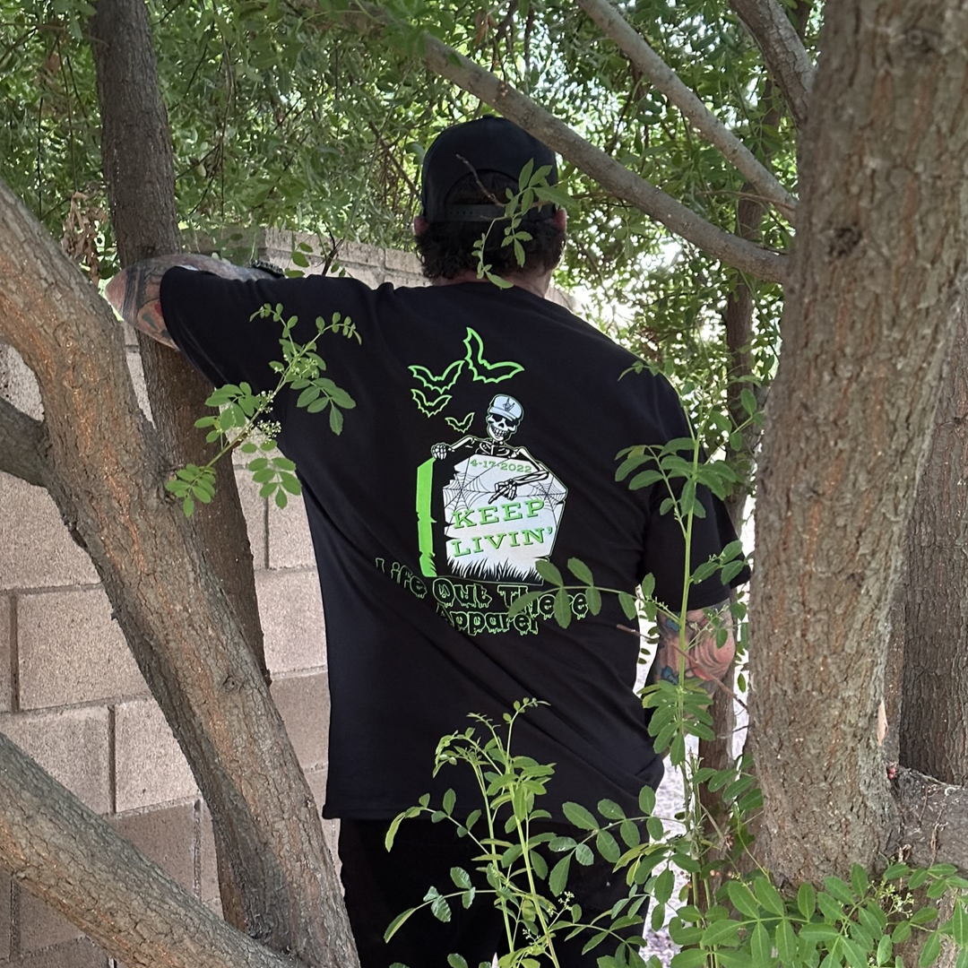 A person dressed in Life Out There Apparel’s black Unisex Keep Livin’ T-Shirt, featuring a skeleton and the "Keep Livin'" design, pairs it with a black cap while standing outdoors near a brick wall surrounded by tree branches, evoking a spooky Halloween atmosphere.